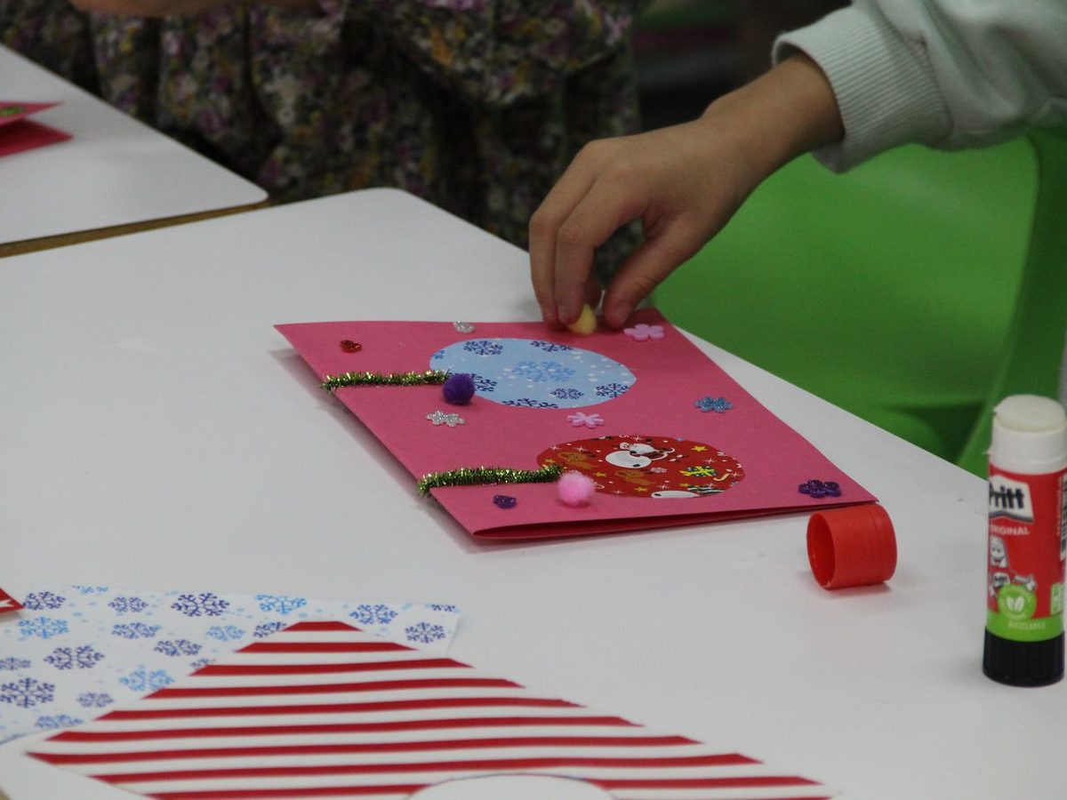 Las manos de un txiki pegan adornos en una tarjeta durante un taller