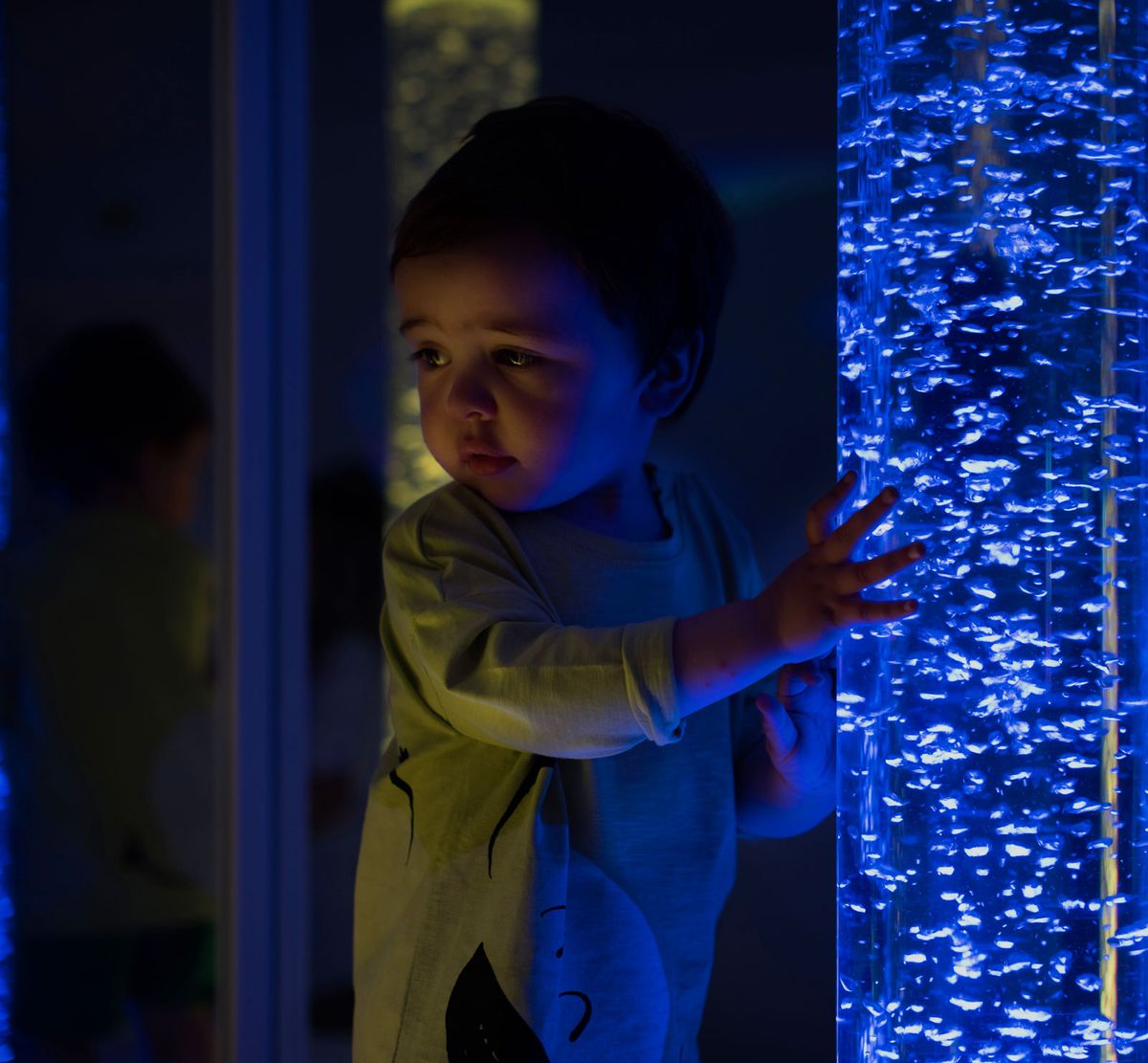 Un niño toca la columna de burbujas del aula Snoezelen, iluminado por su luz azul en la penumbra.