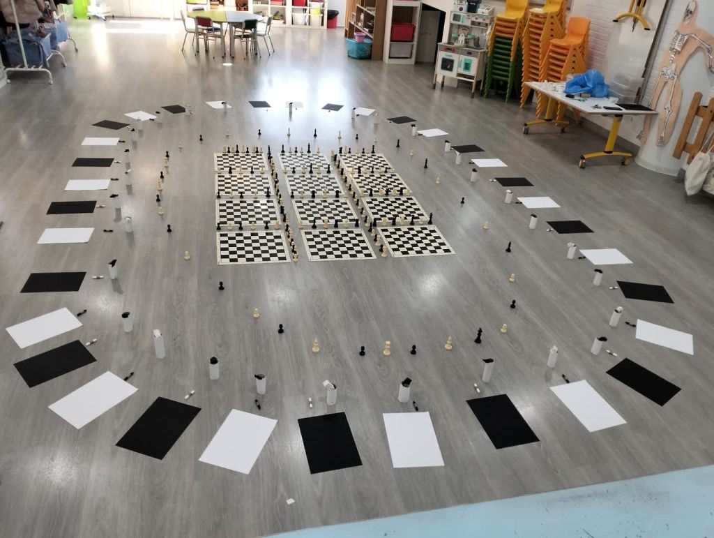 Un tablero de ajedrez gigante montado con piezas y cartulinas en el suelo del aula