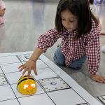 Una niña de infantil coloca un robot con forma de abeja sobre un tapete de cuadrículas